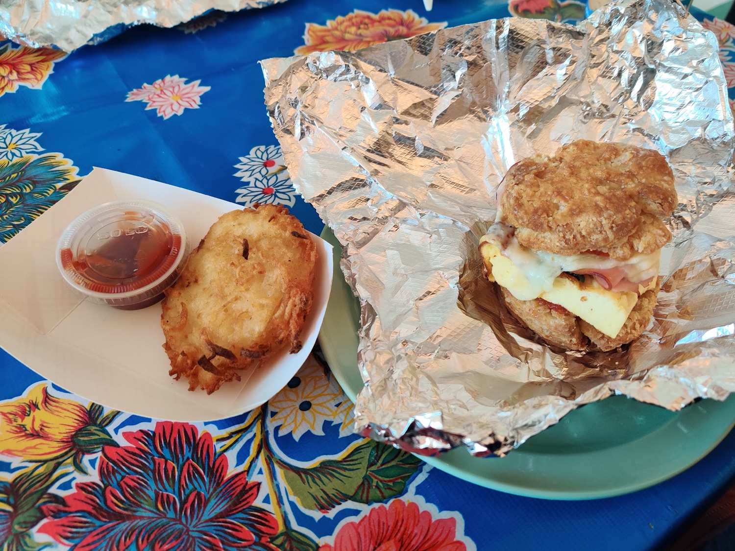 A breakfast sandwich on a buttermilk biscuit, with a hash brown patty and ketchup, on a flowered tablecloth.
