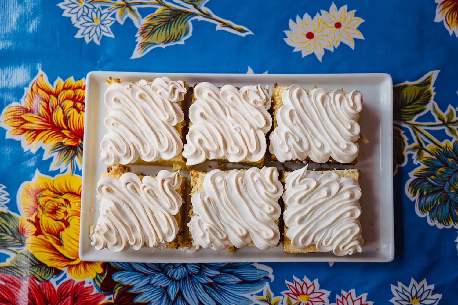 Meringue-topped lemon squares on a white platter, set on a bright floral tablecloth.
